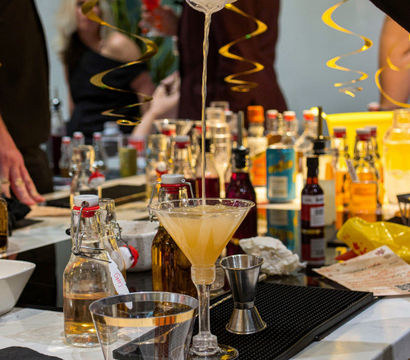 Hand pouring a shaken cocktail into a martini glass at a festive home bar counter with bottles, mixers, and gold streamers.
