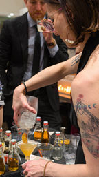Bartender pouring a craft citrus cocktail from a shaker into a martini glass with a dried orange garnish, tattooed arm and hoop earring visible, bottles and glassware on the bar and a suited guest sipping a drink in the background.