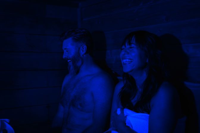 Two adults laughing in a dim, blue-lit wooden sauna — a shirtless man and a towel-wrapped woman seated on a bench, relaxed spa vibe.