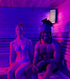 Two people in swimwear sitting on wooden sauna benches under purple-pink neon lighting, smiling next to a wall sconce in a modern indoor sauna.