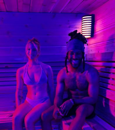 Two people in swimwear sitting on wooden sauna benches under purple-pink neon lighting, smiling next to a wall sconce in a modern indoor sauna.