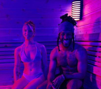 Two people in swimwear sitting on wooden sauna benches under purple-pink neon lighting, smiling next to a wall sconce in a modern indoor sauna.