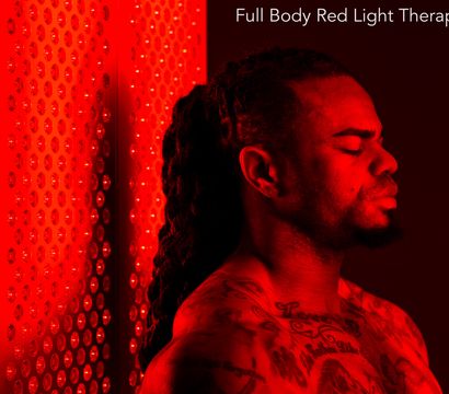 Tattooed man with eyes closed soaking in red LED glow from a full-body red light therapy panel in a wellness studio