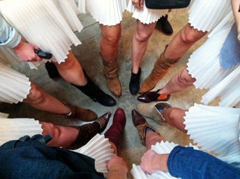 Top-down shot of women in matching white pleated skirts standing in a circle, showcasing a mix of cowboy and ankle boots pointed inward on a concrete floor — playful rustic bridal-party boot photo.