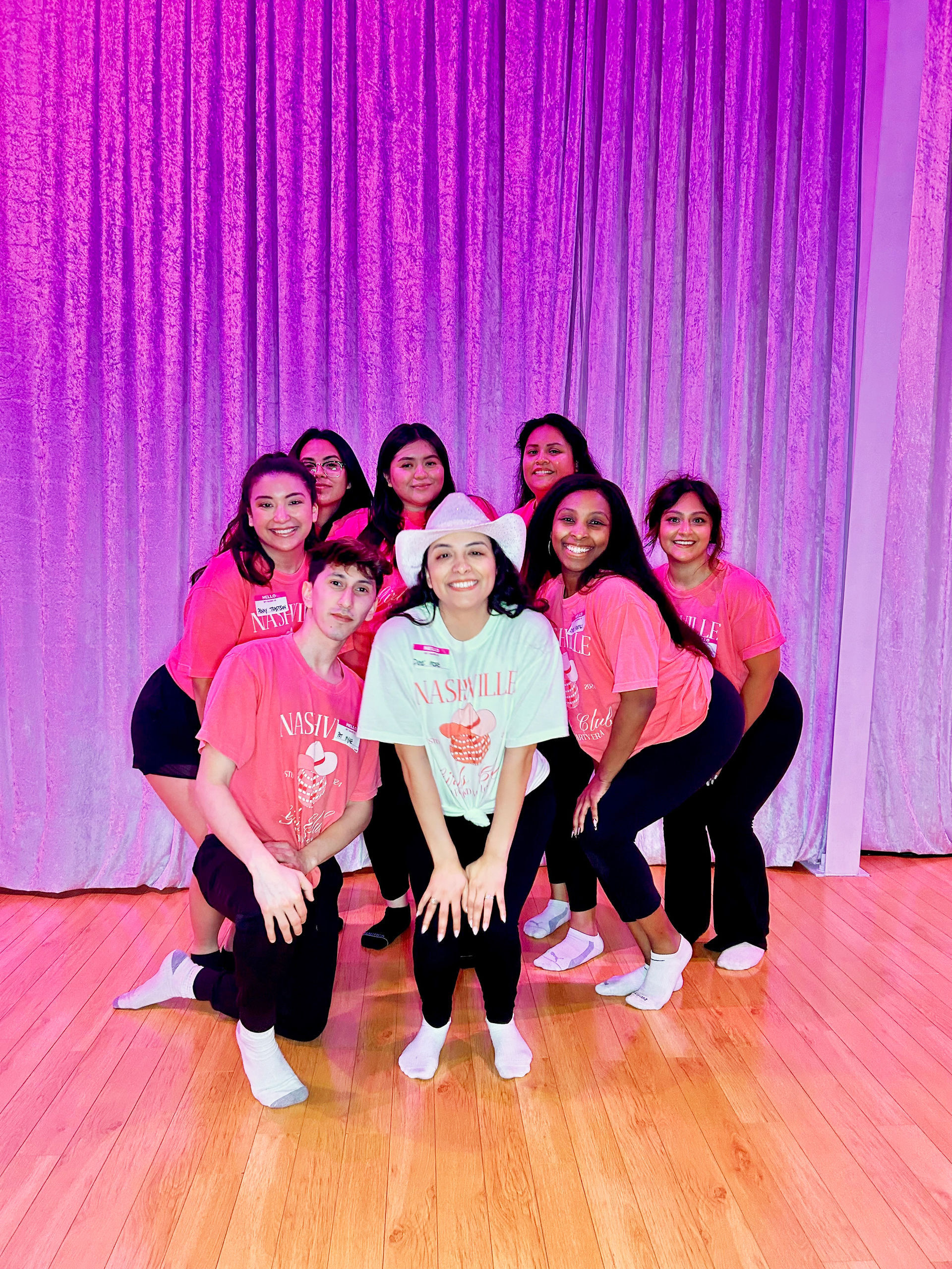 Smiling group of eight young adults in matching pink "Nashville" shirts, one wearing a white cowboy hat, posing on a wooden dance floor in front of purple curtains under pink stage lighting.
