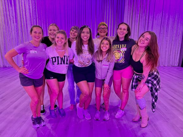 Smiling group of women in activewear posing together in a dance/fitness studio under purple lighting on a light wood floor with curtains in the background.