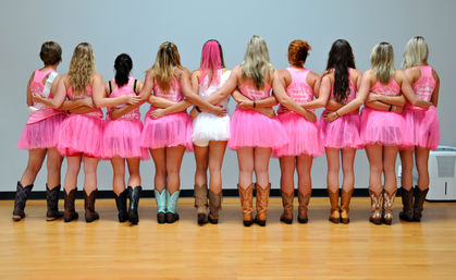 Group of eleven people standing arm-in-arm with backs to camera, wearing bright pink tutus and cowboy boots (one person in white tutu), on a hardwood floor against a plain wall.