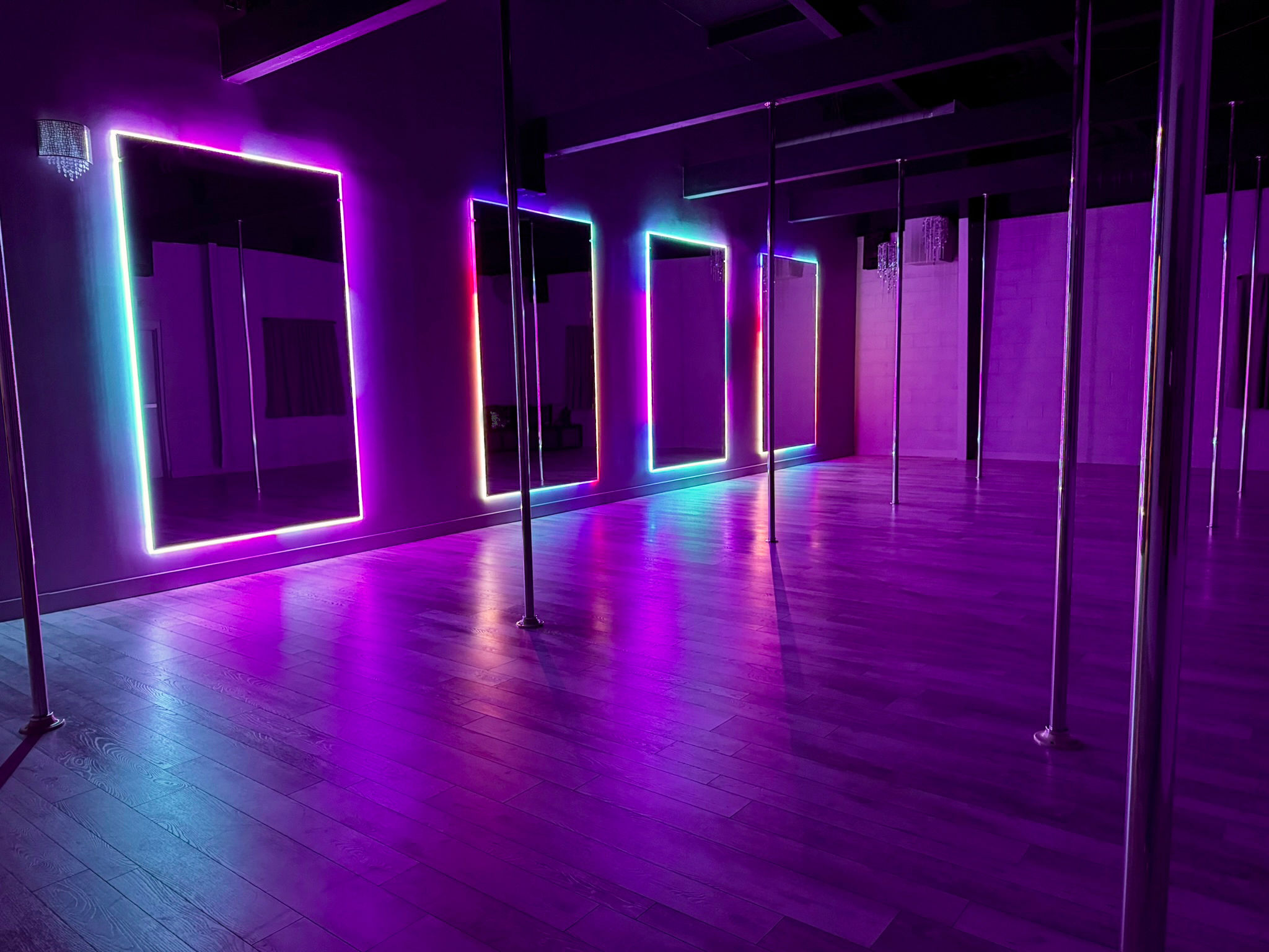 Empty pole fitness studio interior with chrome poles, mirrored walls framed by multicolored LED neon lights and a polished hardwood floor bathed in purple ambient lighting.