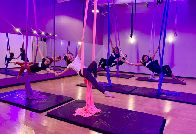 Smiling participants in a group aerial yoga class suspended on pink and blue silks in a purple-lit fitness studio with padded mats and mirrored wall