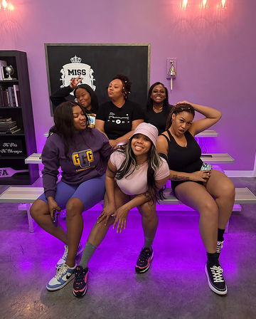 Six women posing playfully on bleacher benches in a pink-and-purple lit studio, wearing casual athletic outfits and sneakers; chalkboard behind reads "MISS" with shelves and a bell visible.