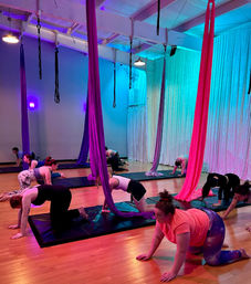 Group aerial yoga class in a neon-lit indoor studio, students on mats practicing tabletop poses beneath hanging magenta, purple and teal aerial silks on a hardwood floor
