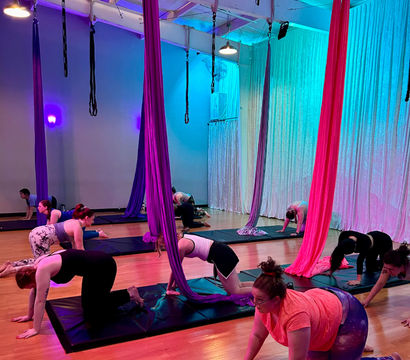 Group aerial yoga class in a neon-lit indoor studio, students on mats practicing tabletop poses beneath hanging magenta, purple and teal aerial silks on a hardwood floor