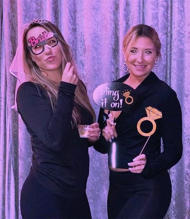Two friends in black outfits pose in front of a purple photo‑booth curtain at a bachelorette party, one wearing a tiara, veil and 'Bride' glasses while holding a drink, the other smiling and holding a large gold ring prop and a 'bling it on!' sign.