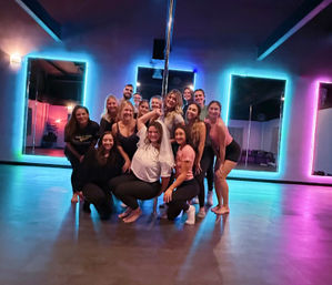 Group of people posing around a chrome pole in a neon-lit pole dance studio with LED-framed mirrors, smiling in workout clothes — upbeat bachelorette party or group pole-fitness class vibe.