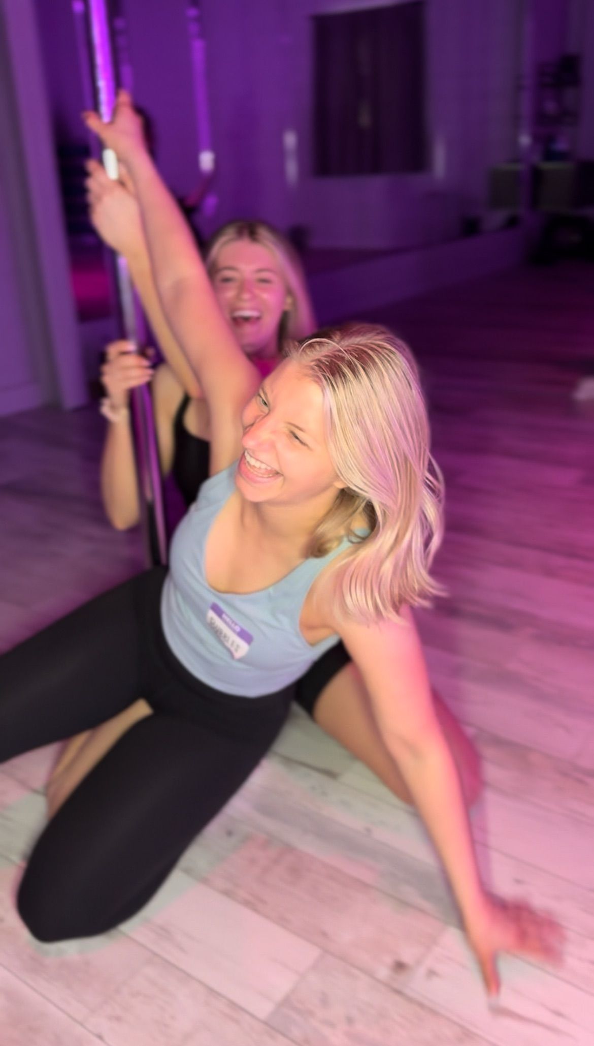Two women laughing during a pole-fitness class in a purple-lit studio, kneeling on light wood flooring.