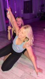 Two women laughing during a pole-fitness class in a purple-lit studio, kneeling on light wood flooring.