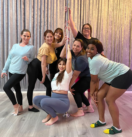 Seven women in workout clothes playfully posing around a chrome pole in a group pole-fitness class, smiling in front of a shimmery curtain and light wood floor.