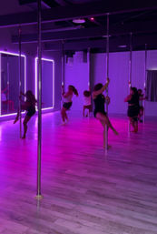 Group pole fitness class with women on chrome poles in a neon purple-lit studio with mirrored wall and light wood floor