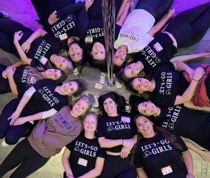 Overhead shot of a circle of smiling women lying feet-out around a chrome pole at an indoor pole-dance party, many wearing matching “LET’S GO GIRLS” shirts with scattered dollar bills and purple mood lighting.