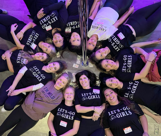 Overhead shot of a circle of smiling women lying feet-out around a chrome pole at an indoor pole-dance party, many wearing matching “LET’S GO GIRLS” shirts with scattered dollar bills and purple mood lighting.