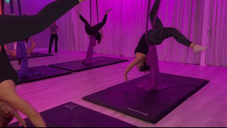 Aerial yoga class in a purple-lit fitness studio with participants suspended upside down in fabric silks above padded mats
