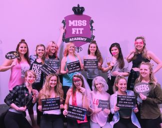 Smiling group of women in a pink-lit fitness studio posing for a bachelorette party, holding novelty props and signs with the bride-to-be wearing a veil.