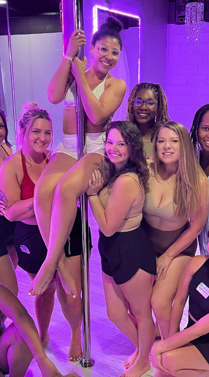 Group of smiling women in activewear posing around a chrome dance pole in a purple-lit pole fitness studio, showing a friendly pole-dancing class atmosphere