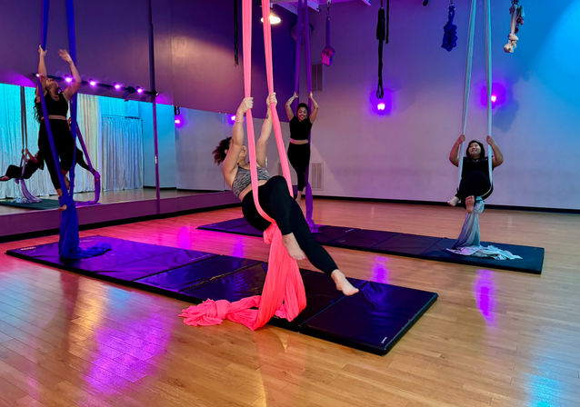 Three participants practicing aerial silks in a fitness studio, suspended on pink, purple and gray silks above padded mats with mirrored wall, hardwood floor and purple-blue mood lighting.