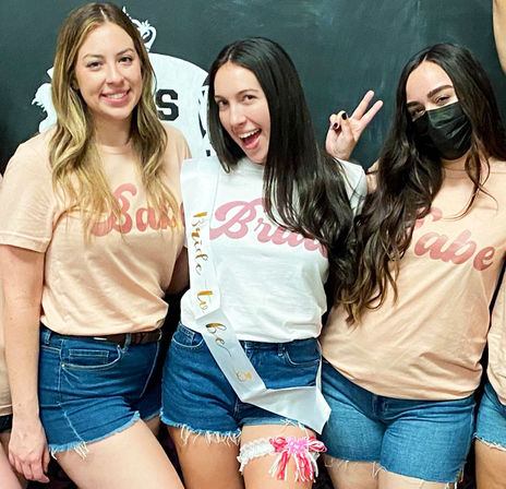 Bachelorette party group photo of three friends in denim shorts and matching pastel t-shirts; center bride-to-be wears a sash and garter, one friend flashes a peace sign and another wears a face mask.