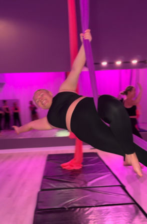Smiling participant in black activewear suspended on purple aerial silks in a pink-lit fitness studio, posing sideways above crash mats