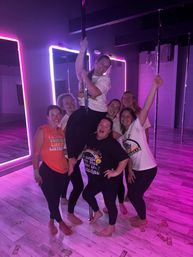 Seven women smiling in a neon‑lit pole dance studio, one climbing a pole while the others pose barefoot on a wood floor scattered with play dollar bills, mirrors and pink‑purple LED lights in the background.