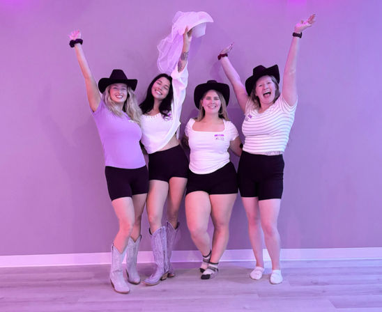 Group bachelorette party photo in a purple-lit studio: four friends wearing cowgirl hats, black shorts and casual tops, raising their arms and holding a bridal veil, one in cowboy boots.