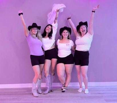 Group bachelorette party photo in a purple-lit studio: four friends wearing cowgirl hats, black shorts and casual tops, raising their arms and holding a bridal veil, one in cowboy boots.
