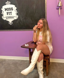 Young woman seated on a vintage wooden school desk against a purple wall, wearing white knee-high cowboy boots and a white tee, playfully licking a lollipop while looking up, with a chalkboard crest and wall bell in the background.