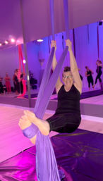 Smiling woman in black workout clothes suspended on purple aerial silks with feet wrapped, hanging over crash mats in a purple-lit aerial yoga/fitness studio with mirrors and classmates in the background.