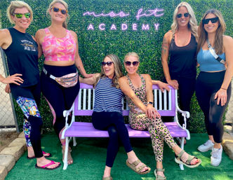 Six smiling women in colorful activewear and sunglasses posing on and around a purple bench against a green hedge backdrop with a neon fitness sign at an outdoor workout event.