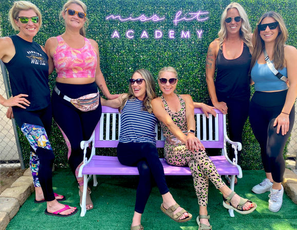 Six smiling women in colorful activewear and sunglasses posing on and around a purple bench against a green hedge backdrop with a neon fitness sign at an outdoor workout event.