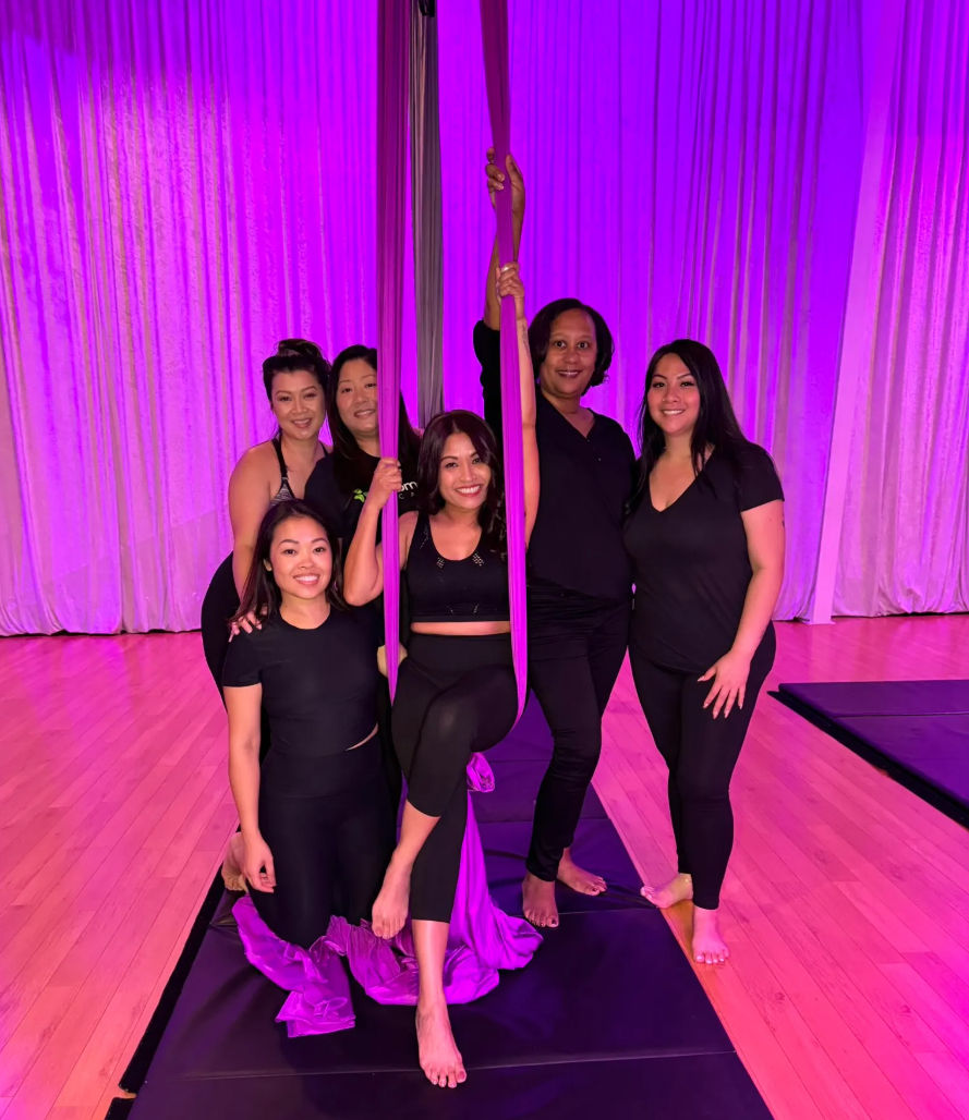 Six women in black activewear smile and pose with purple aerial silks in a brightly lit indoor fitness studio with hardwood floor and draped curtains.