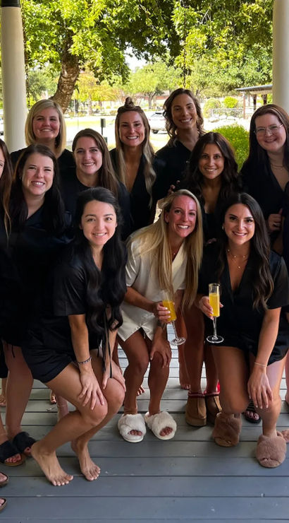 Group of women in matching black and white loungewear and slippers, smiling and holding mimosas on a shaded farmhouse-style front porch with trees and a tree-lined street beyond.