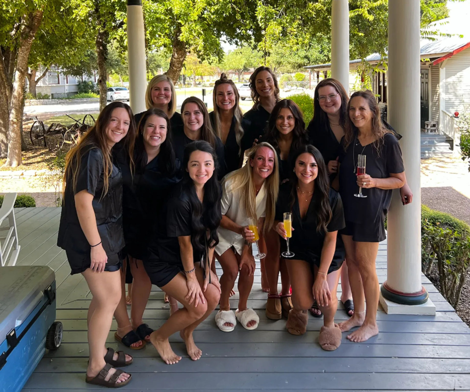 Group of women in matching black and white loungewear and slippers, smiling and holding mimosas on a shaded farmhouse-style front porch with trees and a tree-lined street beyond.
