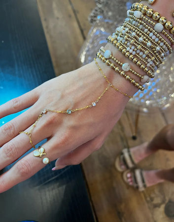 Close-up of a hand wearing stacked gold and white beaded bracelets and a delicate gold hand chain connecting to a pearl butterfly ring, casual wooden-floor background.