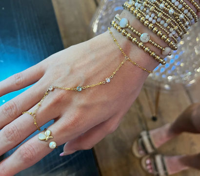 Close-up of a hand wearing stacked gold and white beaded bracelets and a delicate gold hand chain connecting to a pearl butterfly ring, casual wooden-floor background.