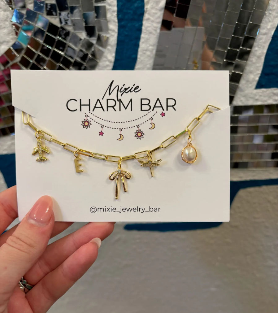 Gold chain charm bracelet on a white display card featuring airplane, letter E, bow, dragonfly and pearl charms, held by a hand with pink nails against mirrored mosaic tiles and a blue wall