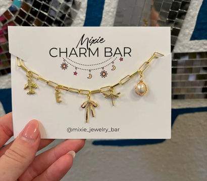 Gold chain charm bracelet on a white display card featuring airplane, letter E, bow, dragonfly and pearl charms, held by a hand with pink nails against mirrored mosaic tiles and a blue wall