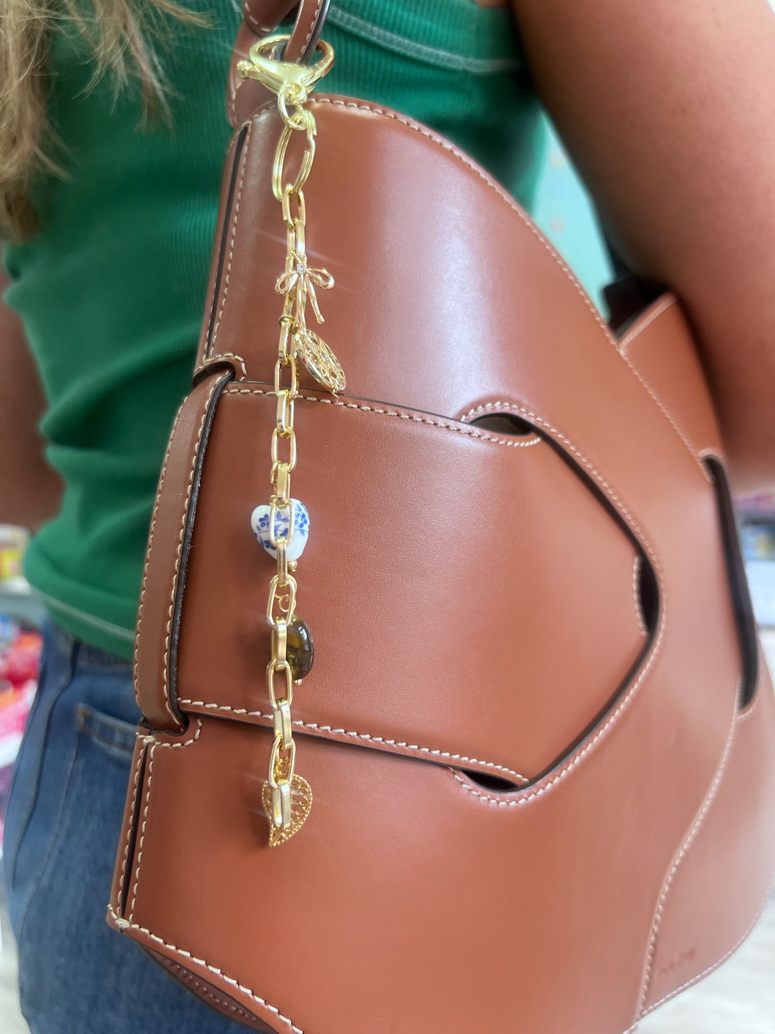 Close-up of a tan brown leather handbag with white stitching and a dangling gold chain charm featuring a blue-and-white bead, tiny gold leaf and heart charms, worn by someone in a green top and jeans.