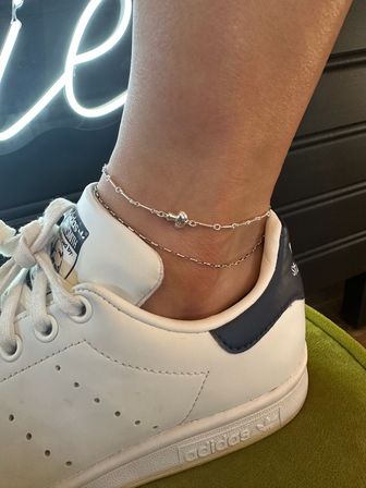 Close-up of an ankle wearing two delicate silver anklets—one with a tiny heart bead—inside a white leather sneaker with a navy heel tab on a green cushion, neon sign glow in the background.