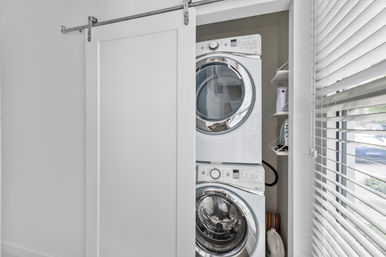 Compact laundry closet with stacked front-loading washer and dryer behind a white sliding barn door, shelf with iron and detergent, and sunlight-filtering window blinds