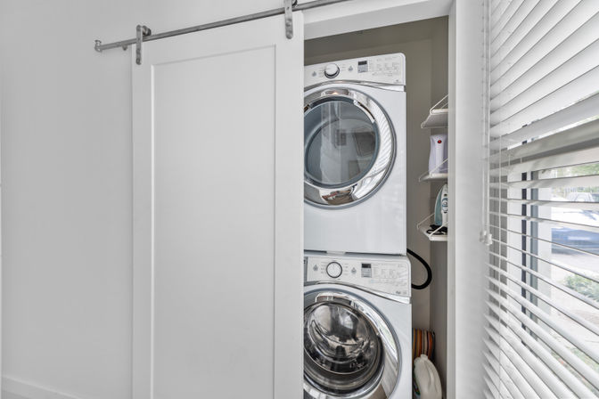 Compact laundry closet with stacked front-loading washer and dryer behind a white sliding barn door, shelf with iron and detergent, and sunlight-filtering window blinds