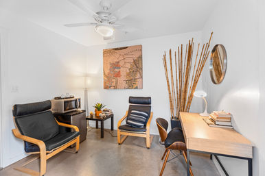Cozy modern home office interior — white walls and ceiling fan, two black leather lounge chairs with wooden frames, small side table with plant and books, wood desk, tall bamboo stalks and round wall mirror.