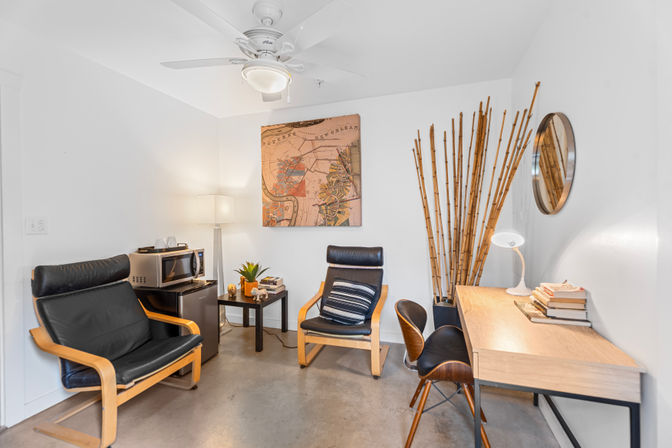 Cozy modern home office interior — white walls and ceiling fan, two black leather lounge chairs with wooden frames, small side table with plant and books, wood desk, tall bamboo stalks and round wall mirror.
